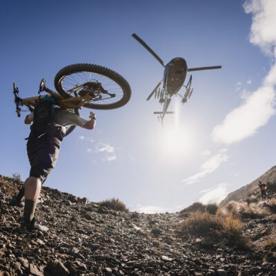 NZMTB Rally - Cam Mackenzie - biker carrying bike uphill with heli over